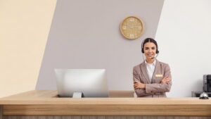 Receptionist wearing a headset standing at a front desk beside a computer and a service bell