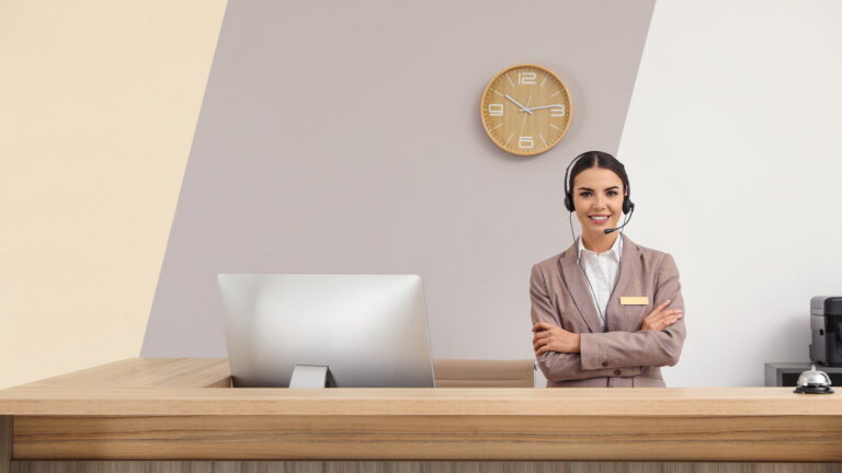 Receptionist wearing a headset standing at a front desk beside a computer and a service bell